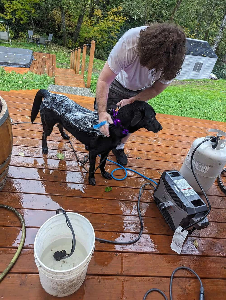 Levi washing his black dog Leo on a wooden deck with a Fogatti portable water heater beside them.