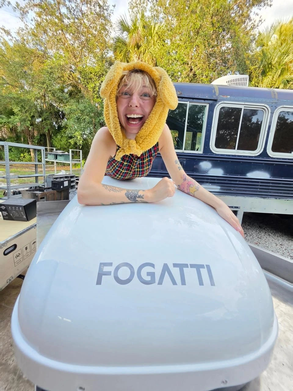 A smiling Busandbunny creator leaning over a white Fogatti air conditioner beside a blue converted bus.